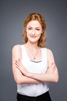 portrait of a young red-haired beautiful girl in the studio on a gray isolated background. A woman is standing with her arms folded and smiling in a white shirt with a short sleeve. Business concept