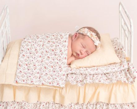 Infant in hairband napping under the sheets of lovely kids' bed with yellowish sheets