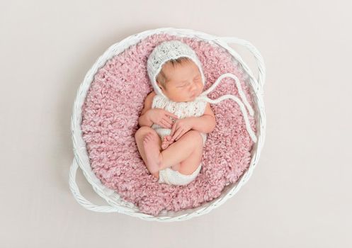 Adorable kid napping on big pink pillow wearing a hat and cute suit, in basket