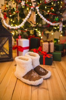 pair of warm home shoes made of wool stands near the Christmas tree with gifts at home on the wooden floor.