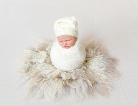 Lovely baby wrapped in white blanket wearing white warm hat sitting in cocoon, sleeping