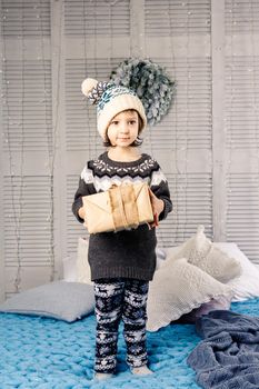 little girl the child sitting in pajamas and hat on the bed with garland of light bulbs with gifts boxes wrapped in a non-colored paper decorated with cones on blue knitted coverlet.Christmas concept.