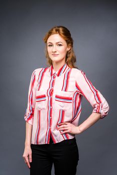 portrait of a young red-haired beautiful girl in the studio on a gray isolated background. A woman stands and smiles in a red shirt with a short sleeve. Business concept.