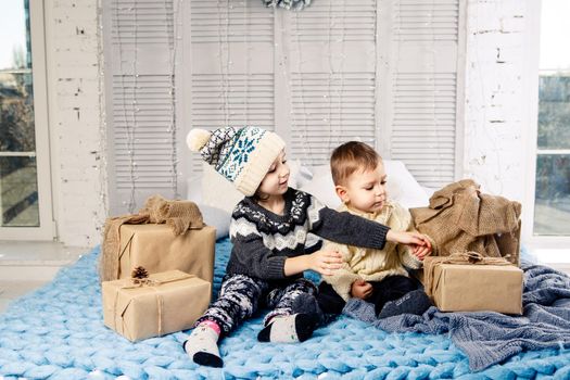 Theme Christmas morning. Two child Caucasian boy and girl brother and sister are sitting on the bed in an embrace with a smile and joy are opening New Year's gifts in boxes on a sunny day.