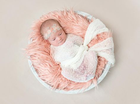 Adorable baby swaddled with white blanket, lying on pink soft pillow, in a basket, topview