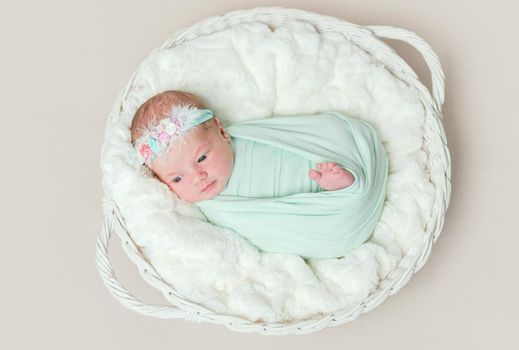 Adorable swaddled baby with legs moving, lying in kids basket, wearing headband