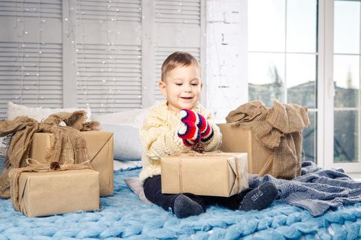 little funny playful boy a child sits on a bed on Christmas day with gift boxes in white wool knitted sweater and big bright mittens on it and laughs out loud. In interior there is a festive decor.