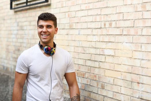 Portrait of young man in urban background smiling with headphones. Wearing white t-shirt near a brick wall