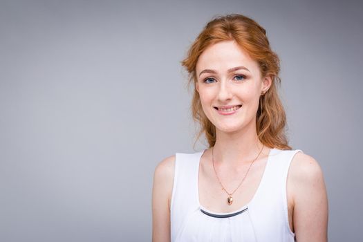 Closeup portrait young, beautiful business woman, student with lred, curly hair and freckles on face on gray background in the studio. Dressed in white blouse with short sleeves about open shoulders.