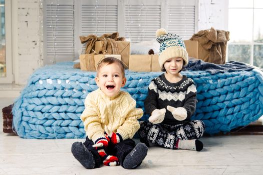 Children brother and sister sit on the floor in bedroom near the bed with gifts on the background of Christmas decor on a sunny day. Dressed in a warm knitted woolen garment cap and colored mittens.