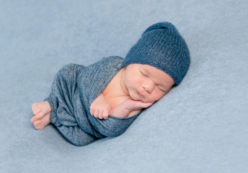 Newborn baby boy swaddled in blue blanket and blue knitted bonnet sleeps peacfully on grey background.