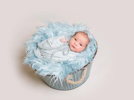 Newborn baby lying on basket covered with blue fluffy rug and curiously looking at camera, top view.