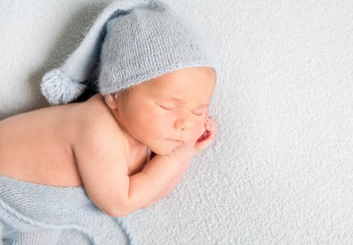 Sleeping baby boy in blue bonnet curled up on white blanket.