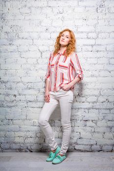 Full-length portrait A young beautiful Caucasian woman student stands near a white brick wall. Girl with red long curly hair with freckles on her face in bright summer clothes.