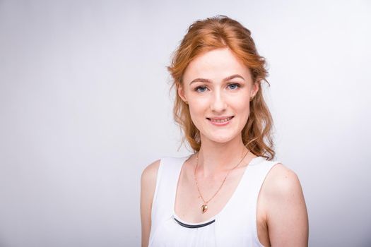 Portrait of a beautiful young female student with a smile in a white shirt of European, Caucasian nationality with long red hair and freckles on her face posing on a white background in the studio.