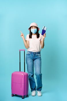 Vertical shot of happy korean woman in medical mask, holding flight tickets and passport, standing with suitcase, dreaming of future vacation, blue background.