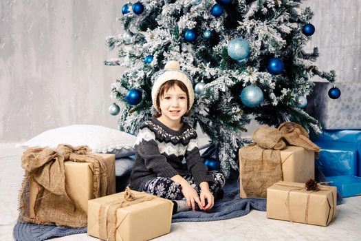 Christmas and New Year concept. A pretty girl in a white knitted hat and sweater sits on the floor near a gift box on the background of a beautiful Christmas tree.
