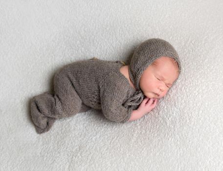 Dreamy baby boy wearing brown knitted clothes sleeps on his side on white background.