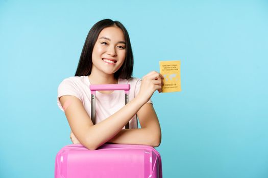 Happy smiling asian girl, tourist stands with cute suitcase in airport, shows international covid-19 vaccination certificate for travellers, travelling safe after vaccination, blue background.