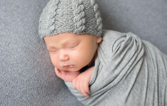 Beautiful newborn baby sleeping curled up in grey blanket and wearing grey hat. Little baby lying on grey surface