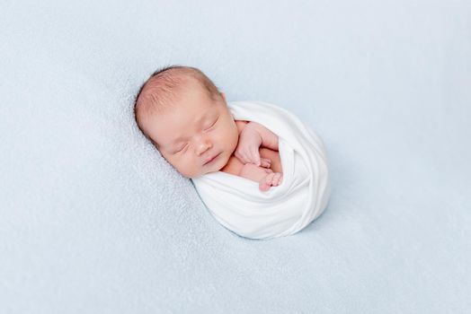 Little newborn baby sleeping swaddled in a white blanket on blue background