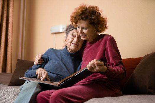 Mature daughter visits senior mother and has fun watching family photo album, sitting in embrace in living room couch. Mothers Day. Elderly woman and old gray hair grandmother laugh and remember youth