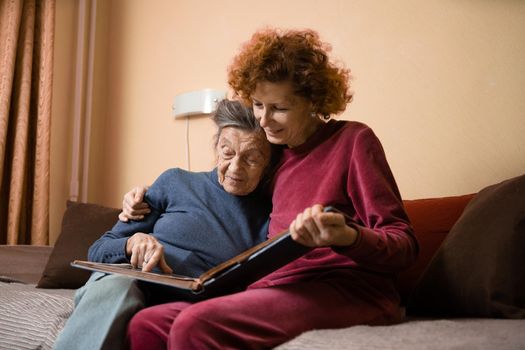 Senior woman and her adult daughter looking at photo album together on couch in living room, talking joyful discussing memories. Weekend with parents, family day, thanksgiving, mom's holiday.