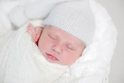 Top view of newborn baby wrapped in white wrap wearing white bonnet and lying on white round basket with soft blanket. Newborn baby in white outfit