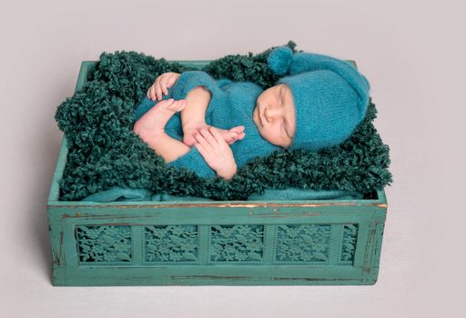 Beautiful newborn baby in green cap lying in wooden crate with arms under the head