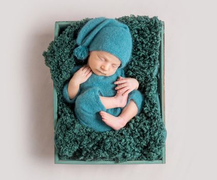 Beautiful newborn baby in green cap lying in wooden crate with arms under the head