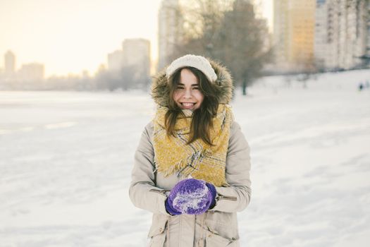 Portrait of a beautiful young Caucasian woman in a knitted hat and scarf standing on a winter background with snow smile and happiness purple gloves sculpt a snowball.
