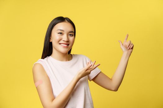 Covid-19 vaccination concept. Smiling asian vaccinated girl with patch on shoulder, pointing fingers at healthcare banner, yellow background.