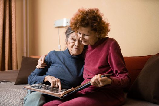 Positive aged ladies looking album photos sitting sofa at home, cheerful friends. Senior woman and her mature nurse watching photo album. Granny showing her daughter memories from the past.