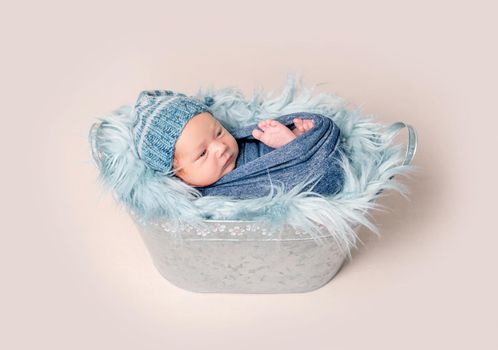 Newborn baby lying in trough with blue fluffy blanket. Little newborn baby boy wrapped in blue blanket and wearing in blue bonnet