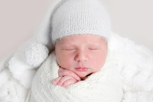 Top view of newborn baby wrapped in white wrap wearing white bonnet and lying on white round basket with soft blanket. Newborn baby in white outfit