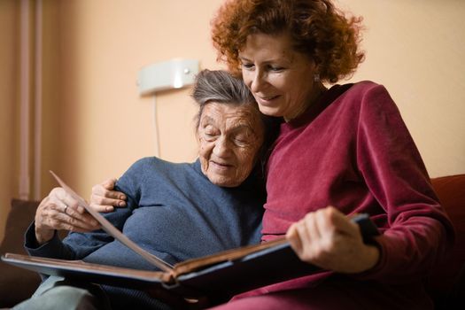 Mature daughter visits senior mother and has fun watching family photo album, sitting in embrace in living room couch. Mothers Day. Elderly woman and old gray hair grandmother laugh and remember youth