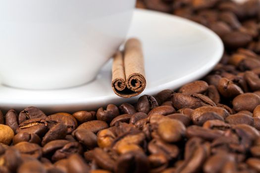 White cup on a pile of coffee beans, on a plate lie whole cinnamon sticks