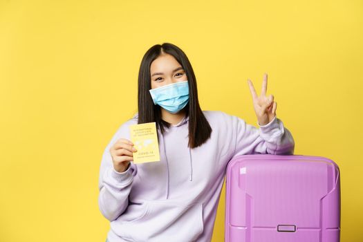 Smiling asian girl tourist in face mask, standing with suitcase, showing covid international vaccination certificate for travellers and peace sign, yellow background.