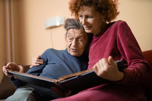 Positive aged ladies looking album photos sitting sofa at home, cheerful friends. Senior woman and her mature nurse watching photo album. Granny showing her daughter memories from the past.