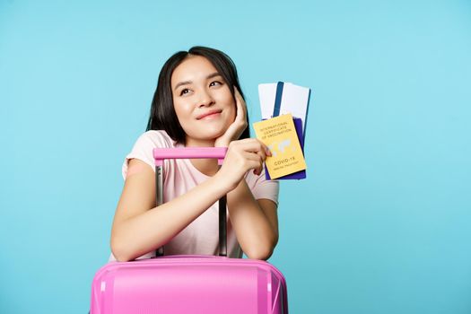 Dreamy asian tourist, smiling and thinking, holding covid-19 international vaccination certificate, two tickets and passport, standing with suitcare for trip, blue background.