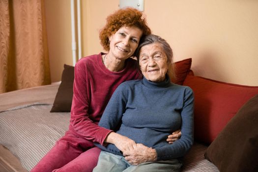 Mother and daughter, mature woman with red hair and elderly old cute happy lady woman with gray hair and deep wrinkles, embrace together at home and smile happily, family ties and caring for parents.