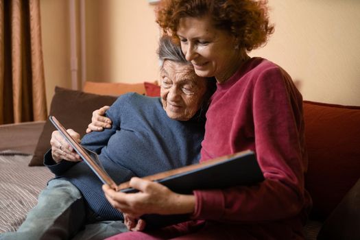 Senior woman and her adult daughter looking at photo album together on couch in living room, talking joyful discussing memories. Weekend with parents, family day, thanksgiving, mom's holiday.