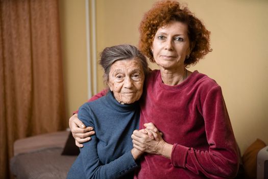 Mother and daughter, mature woman with red hair and elderly old cute happy lady woman with gray hair and deep wrinkles, embrace together at home and smile happily, family ties and caring for parents.