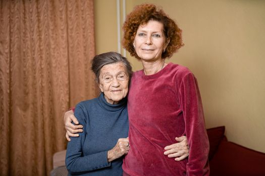 Senior Caucasian woman tenderly hugs her ninety year old elderly mother with gray hair, face with deep wrinkles, at home on sofa, smiling mother and daughter. Theme care and dedication to old parents.
