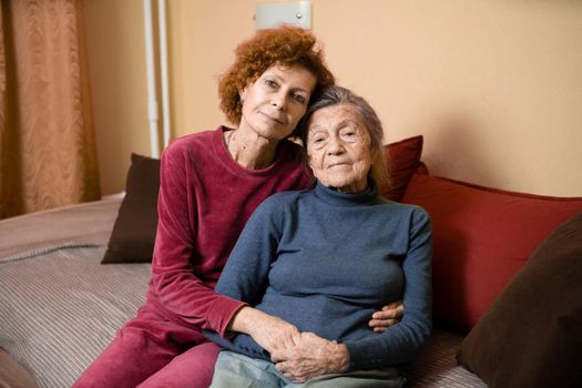 Mother and daughter, mature woman with red hair and elderly old cute happy lady woman with gray hair and deep wrinkles, embrace together at home and smile happily, family ties and caring for parents.