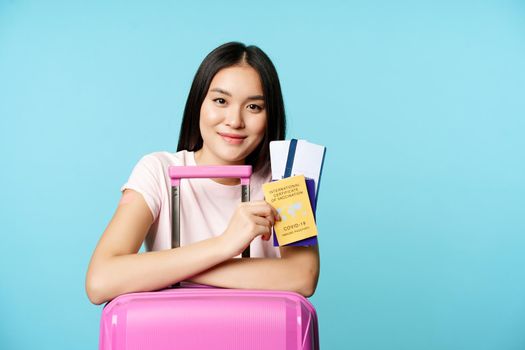 Happy asian girl with suitcare, shows coronavirus international vaccination certificate, two tickets for travelling abroad, going on trip after covid-19 vaccine, blue background.