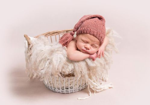 Little baby in light pink-red knitted beanie sweetly sleeping on the white coverlet in the basket on the light background