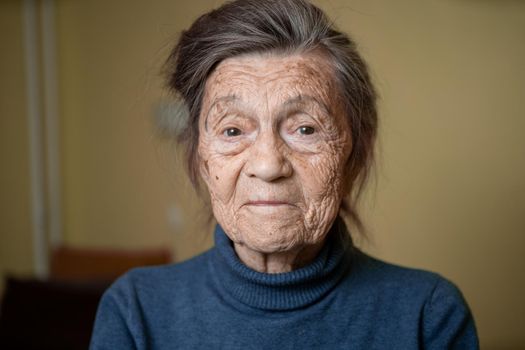 90 year old cute elderly woman with gray hair and wrinkles face, wearing sweater, portrait large, smiling and looking joyfully, background of room. Theme long-liver and aging, old people in good mood.