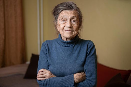 90 year old cute elderly woman with gray hair and wrinkles face, wearing sweater, portrait large, smiling and looking joyfully, background of room. Theme long-liver and aging, old people in good mood.