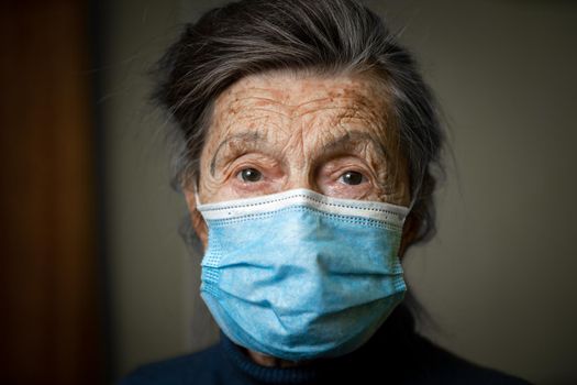 Elderly Caucasian woman with wrinkles on face, gray hair, wearing medical mask, looks attentively at camera, feeling need care and support. Topic safe communication, stay at home during diseases.